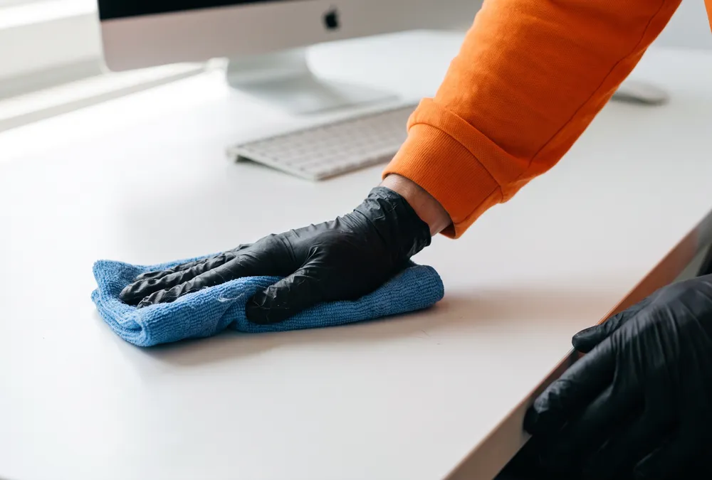 Main gantée essuyant un bureau blanc avec une microfibre bleue, devant un écran d'ordinateur